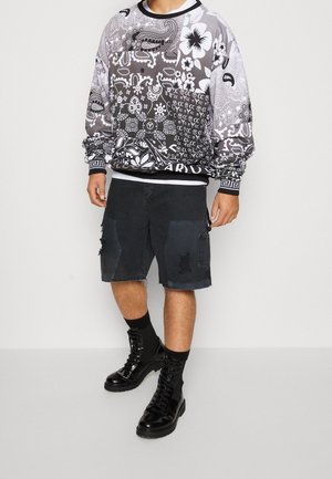 Schwarz-weiß gemustertes Sweatshirt mit floralen und Paisley-Designs, kombiniert mit schwarzen Cargo-Shorts mit ausgefranstem Saum und Rissen. Schwarze Kampfstiefel.