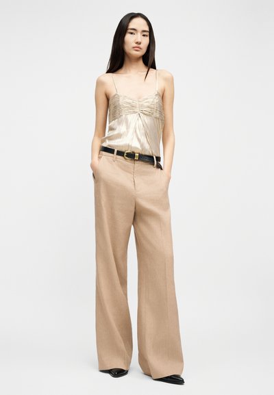 Νεαρή γυναίκα με μακριά σκούρα μαλλιά, ντυμένη με μπεζ σατέν καμιζόλ, παντελόνι σε ταμπά χρώμα με φαρδιά πόδια, μαύρη ζώνη και μαύρα μυτερά παπούτσια, όρθια με τα χέρια στις τσέπες.