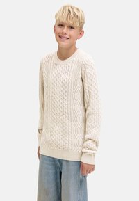 Blonder Junge, der einen cremefarbenen kabelgezopften Pullover und hellblaue Jeans trägt, lächelt und steht vor einem schlichten Hintergrund.