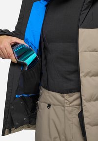 Schwarze und beige isolierte Jacke mit blauer Innenauskleidung, ausgestattet mit einer Netztasche, die mehrfarbige Skibrillen hält. Texturiertes Material und Druckknöpfe sind sichtbar.