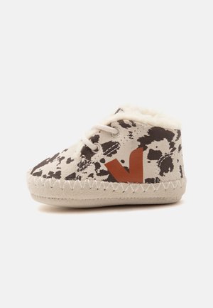 Chausson bébé doux avec imprimé vache noir et crème, lacets blancs, logo marron « "V" » et doublure blanche duveteuse sur semelle beige.