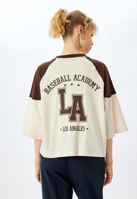 Camiseta de béisbol de color marrón y crema con mangas cortas. Presenta "BASEBALL ACADEMY LA" impreso en la parte posterior en letras marrones oscuras.