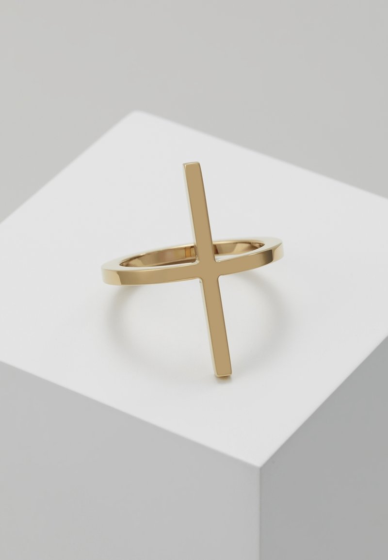 Bague en or avec une fine bande présentant une forme de croix verticale, posée sur une surface géométrique blanche avec un fond gris neutre.