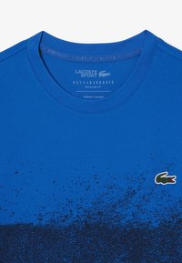 Camisa deportiva azul con textura degradada, que presenta un logo en la esquina inferior derecha y texto en el área del cuello. Diseño de corte regular.