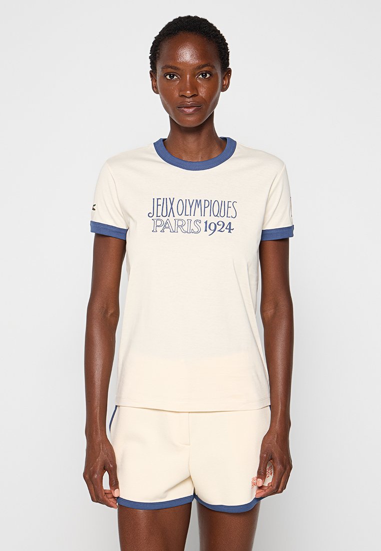 Cremefarbenes T-Shirt mit blauen Rippkragen und Ärmeln. Bedruckt mit dem Text "JEUX OLYMPIQUES PARIS 1924" in Marineblau. Aus weichem, glattem Baumwollmaterial.