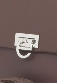 Bruine leren handtas met een zilverkleurige metalen sluiting met een cirkelontwerp en rechthoekige basis. Textuur met minimale details.