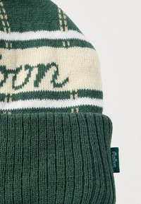 Πλεκτός σκούφος σε σκούρο πράσινο και κρέμ χρώμα, με ριμπ υφή, οριζόντιες ρίγες και διακοσμητικό κείμενο σε καλλιγραφικό στυλ. Περιλαμβάνεται μικρή ετικέτα με το λογότυπο.