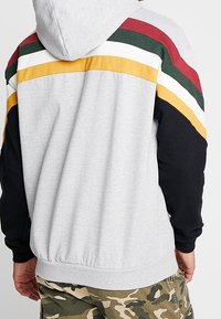 Hombre con sudadera gris con rayas rojas, verdes, blancas y amarillas en los hombros, combinado con pantalones de camuflaje, visto desde atrás.