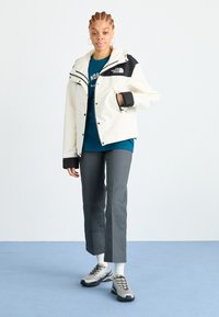 White jacket with black accents, paired with a teal shirt and gray trousers. Model wears gray sneakers with black detailing and white socks.