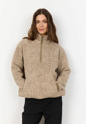 Pullover in pile color sabbia con colletto alto, zip a quarto davanti e tasche laterali. Presenta una superficie morbida e testurizzata e un bordo beige a contrasto.