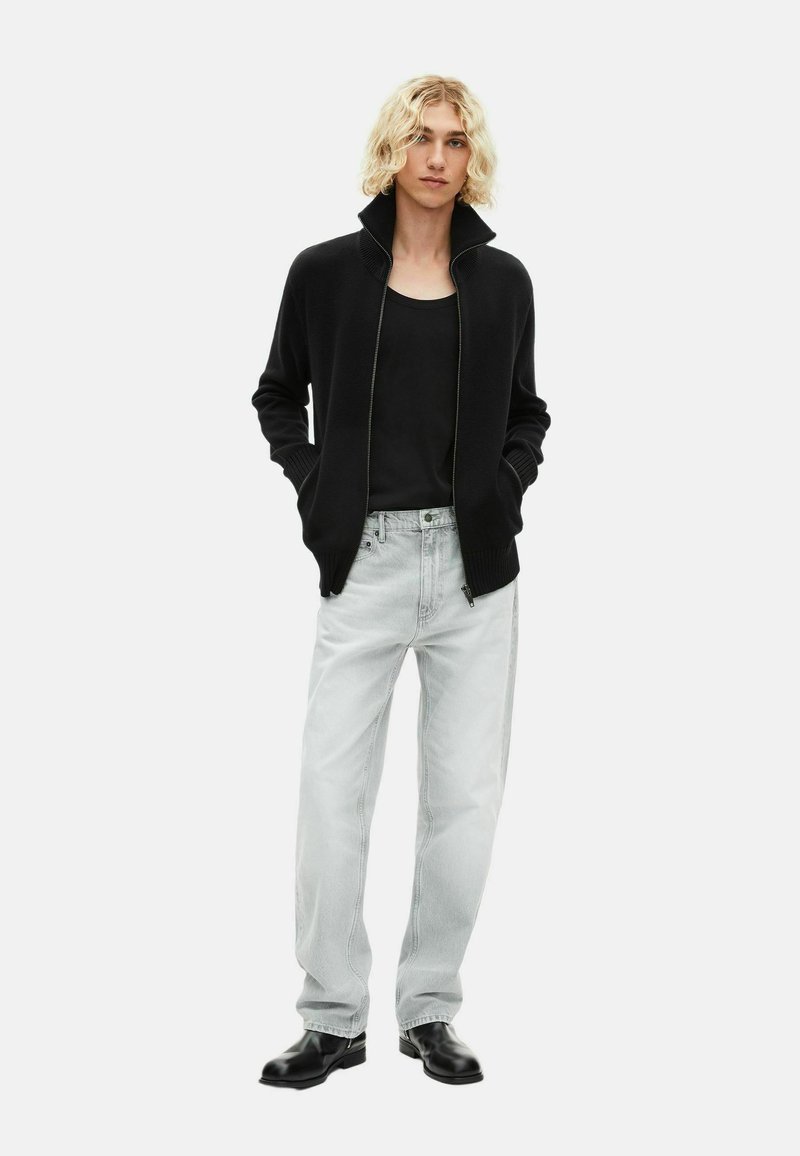 Junge Person mit lockigem blonden Haar, die einen schwarzen Reißverschluss-Pullover, ein schwarzes Oberteil, hellgraue Jeans und schwarze Schuhe trägt. Sie steht mit den Händen in den Taschen.