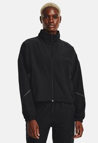Giacca cropped nera con colletto alto, chiusura con zip e polsini elasticizzati; presenta dettagli riflettenti sulle maniche e una texture liscia.