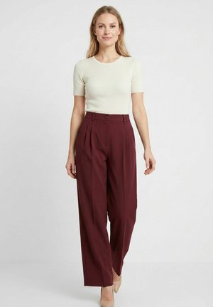 Femeie stă în picioare pe un fundal simplu purtând un top crem cu mâneci scurte, tricotat cu reieșituri, pantaloni burgundy cu talie înaltă și croială largă și pantofi cu toc bej.