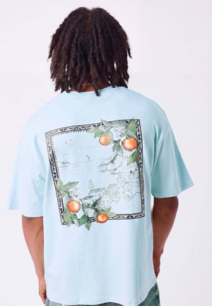 Personne avec des dreadlocks portant un t-shirt bleu clair avec une illustration d'un village côtier pittoresque et un fruit orange au dos.