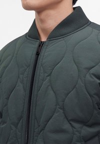 Gros plan d'une personne portant une veste matelassée vert foncé avec un col côtelé et une fermeture éclair noire à l'avant fermée.