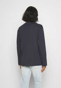 Person med axellångt mörkt hår som bär en marinblå långärmad skjorta och ljusblå jeans, stående framåtvänd mot en enfärgad vit bakgrund.