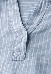 Tessuto a righe blu chiaro con una texture morbida. Presenta un collo a V, una piccola chiusura con bottone e dettagli di cucitura a vista.