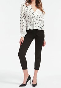Kvinna iförd en vit långärmad blus med svarta prickar, knuten i midjan, svarta slim-fit capribyxor och svarta högklackade skor.