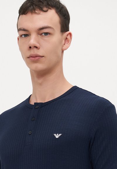 T-shirt henley côtelé navy avec trois boutons, arborant un petit logo blanc sur la zone de la poitrine gauche, fabriqué à partir d'un tissu doux.