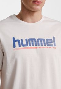 Ljus beige bomullströja med en blå och röd "hummel PERFORMANCE" logotyp, med texturerad skrift och korta ärmar.