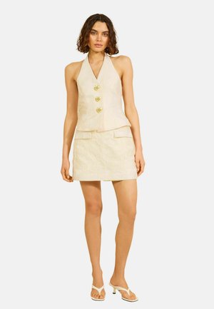 Kobieta ubrana w kremową kamizelkę na szyję z złotymi guzikami i pasującą mini spódniczkę, stoi w białych, otwartych na palce sandałach na obcasie na białym tle.