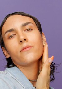 Primo piano di una persona con capelli corti e scuri, che indossa una giacca di jeans e si tocca il collo. La pelle è liscia con un finish naturale. Sfondo viola.