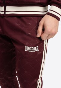 Bordo trenirka s bijelim prugama. Ima suptilan uzorak životinja i istaknuti Lonsdale London logo na nogavici. Mekani materijal.