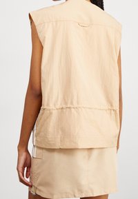 Beige ärmlös topp med en texturerad yta, med en avslappnad passform, en bakficka och vita sidostrickor, ihop med en matchande kort kjol.