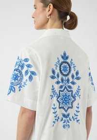 Hvit kortermet skjorte med intrikat blå og brun blomsterbroderi på ryggen og ermene, laget av lett stoff.