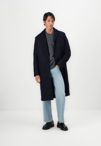 Cappotto di lana navy con colletto a punta, bottoni frontali e tasche laterali, indossato sopra un maglione grigio e jeans azzurri chiari. Scarpe nere.
