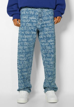 Blauwe spijkerbroek met witte graffiti-stijl tekst over het hele oppervlak, gedragen met witte sneakers en een blauwe sweater.