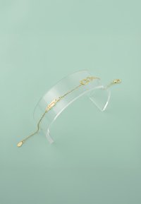 Guldarmband med en fjäderdetalj, oändlighetssymbol och droppformad berlock på en fin kedja, utställt på en klar akrylställ.