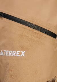 Vodootporna bež tkanina s crnim detaljima na patentnom zatvaraču, koja sadrži kapljice vode. Bijeli logo "TERREX" istaknut je na vidnom mjestu.