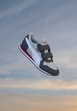 Basket Puma grise et blanche avec des accents rouges et deux bandes velcro flottant sur un fond doux de ciel bleu et beige.