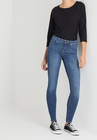 Dunkelblaue Skinny-Jeans aus Denim mit klassischem Fünf-Taschen-Design und leichtem Verblassen, kombiniert mit einem schwarzen Langarmoberteil und schwarzen Sneakers.