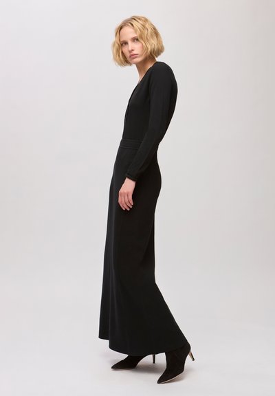 Femme aux cheveux blonds courts portant une longue robe noire et des bottines noires à talons, debout devant un fond clair uni.