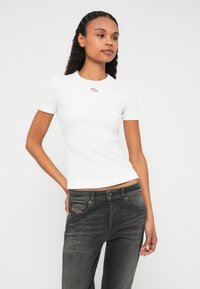 Biele priliehavé tričko s krátkymi rukávmi s rebrovanou textúrou a malým kovovým logom na hrudi, v kombinácii so sivými denimovými džínsami.