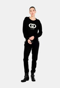 Felpa nera con logo LIU-JO, abbinata a pantaloni jogger neri con dettagli in borchie. Modella indossa stivaletti neri con cinturini decorativi.