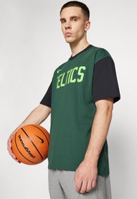 Camisa de manga corta verde y negra con la palabra "CELTICS" impresa en verde, combinada con pantalones de chándal grises. Balón de baloncesto naranja con detalles en negro sostenido en una mano.
