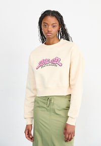 Felpa beige tagliata con logo rosa "Santa Cruz Skateboards", polsini a coste e vestibilità rilassata, abbinata a una gonna verde.
