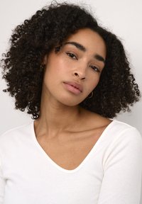 Witte geribde langemouwen top met een V-hals, gedragen door een persoon met donker, krullend haar, blootgestelde schouders en minimale make-up.