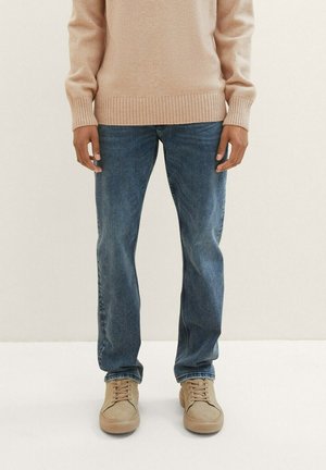 Beige crewneck-neule yhdistetty sinisiin farkkuihin ja vaaleisiin nauhallisiin kenkiin. Rentoa istuvuutta, tavalliset hihansuut ja farkuissa on hieman kulumista.