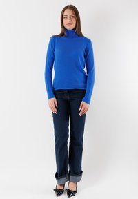 Maglione a collo alto blu dal design aderente, abbinato a jeans blu scuro ripiegati ai polsini, e scarpe nere verniciate con fiocchi.
