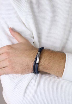 Geflochtenes Armband aus navyblauem Leder mit einem silbernen Metallakzent, getragen an einem Handgelenk, vor einem strukturierten weißen Stoffhintergrund.