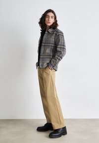 Veste-chemise à carreaux gris et beige, coupe ample, à boutons, portée sur un t-shirt noir et associée à un pantalon large beige et des bottes noires.