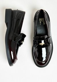 Mocasines de charol negro con un acabado brillante, que presentan una correa decorativa y herrajes en tono dorado. Tacón bajo de bloque, textura suave.