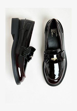 Fekete lakkbőr mokaszinok fényes felülettel, dekoratív pánttal és aranyszínű fémszerelvényekkel. Alacsony tömbsarok, sima textúra.