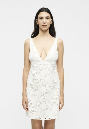 Γυναίκα με μακριά σκούρα μαλλιά, που φοράει ένα λευκό δαντελένιο φόρεμα χωρίς μανίκια με βαθύ V-λαιμόκοψη, στέκεται μπροστά σε ένα απλό ανοιχτό φόντο.