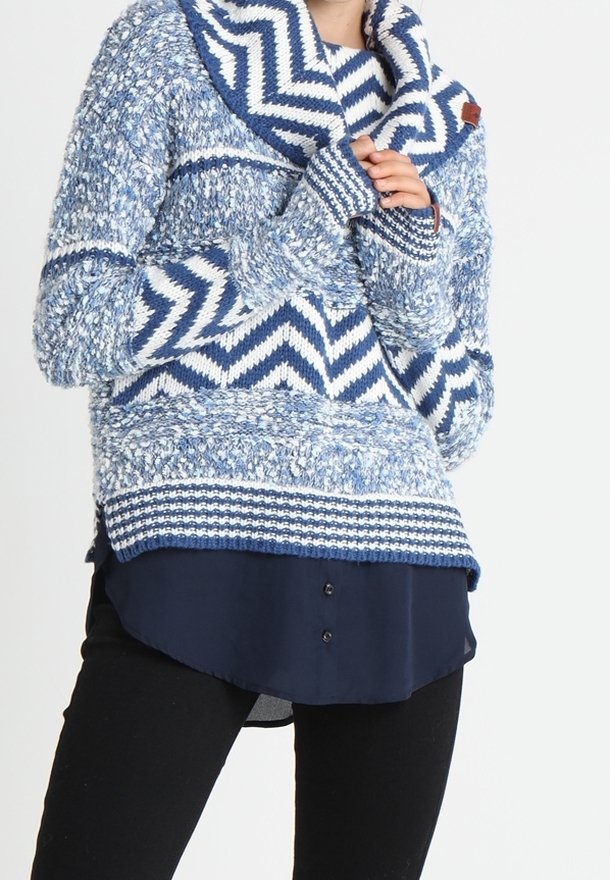 Un pull en tricot bleu et blanc à motifs avec un col drapé, présentant un design en zigzag, un tissu texturé et des accents côtelés. Porté sur une chemise sombre.