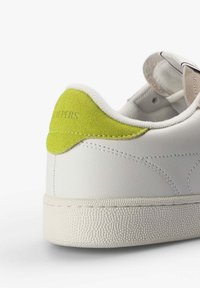 Zapatilla blanca con suela texturizada y parche de ante verde lima en el talón, mostrando costuras detalladas y un cuello acolchado.